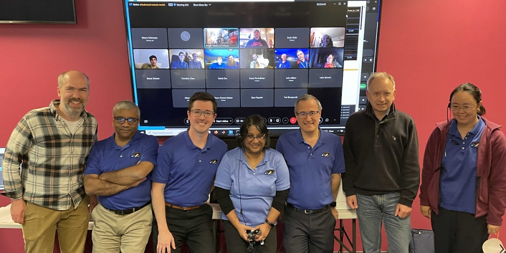 Mitglieder des JPL-Teams posieren, nachdem das erste gestreamte Ultra-HD-Video aus dem Weltraum empfangen wurde. Ferngesteuerte Teammitglieder (einschließlich Taters, der Katze) erscheinen auf dem Konferenzbildschirm. Stehend, von links, sind: Dan Goods, Abi Biswas, Ryan Rogalin, Meera Srinivasan, Bill Klipstein, Oliver Lay und Christine Chen.