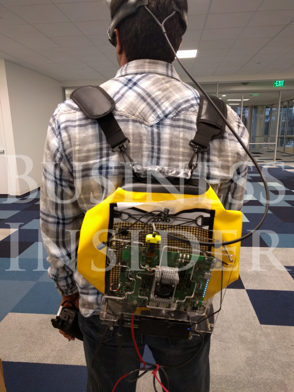 Der Prototyp wird wie ein Rucksack getragen. Die nackten Platinen lassen deutlich erkennen, dass das Gerät in einem frühen Entwicklungsstadium ist. In der linken Hand hält die Person etwas, das der Akku des Gerätes sein könnte. Über die Brille lässt sich nicht viel sagen.