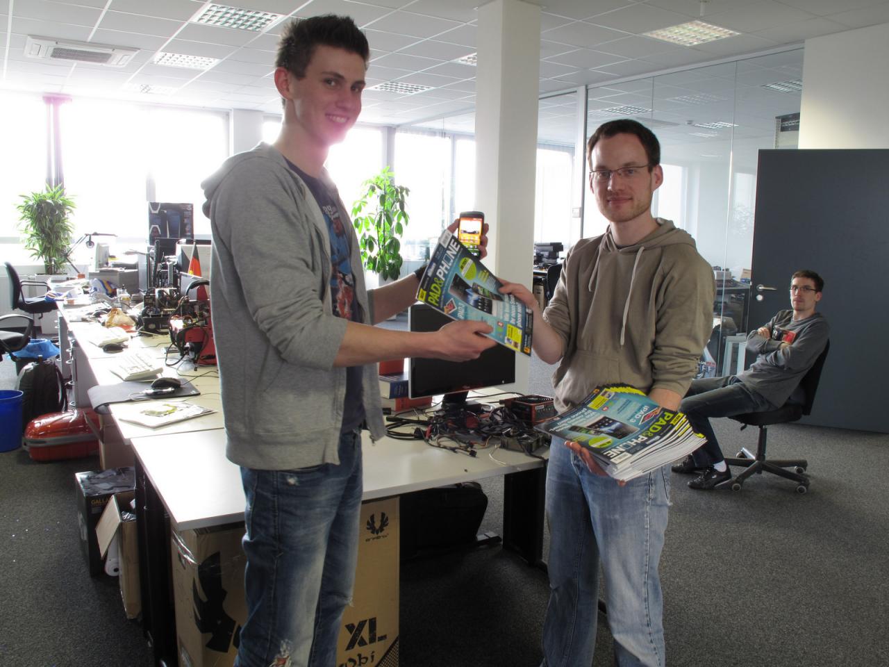 [30/03/2012] Nein, ein Zeitungsverkäufer ist Daniel sicherlich nicht, trotzdem überreicht er voller Stolz eine der neue Pad&Phone-Ausgaben an unseren Fabian, der voller Testeifer schon gleich wieder mit einem Smartphone anrückt, das in der nächste Ausgabe getestet werden könnt. Hoffentlich denkt Daniel beim Verteilen seines Meisterwerks auch an den Clemens im Hintergrund, der sicher auch ein Exemplar haben möchte. 