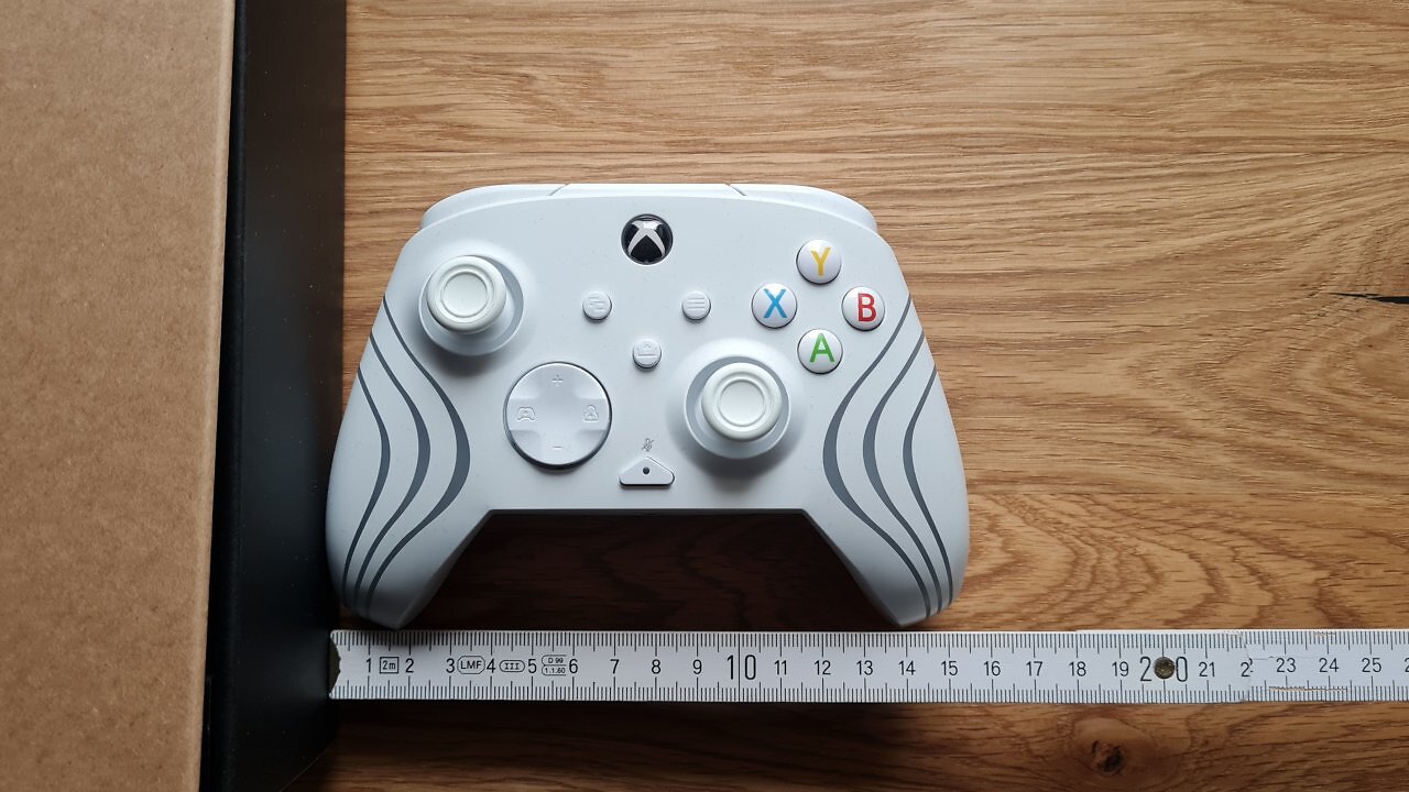 Ein weißer Controller auf einem Holztisch, darunter ein Meterstab, der die Breite zeigt.
