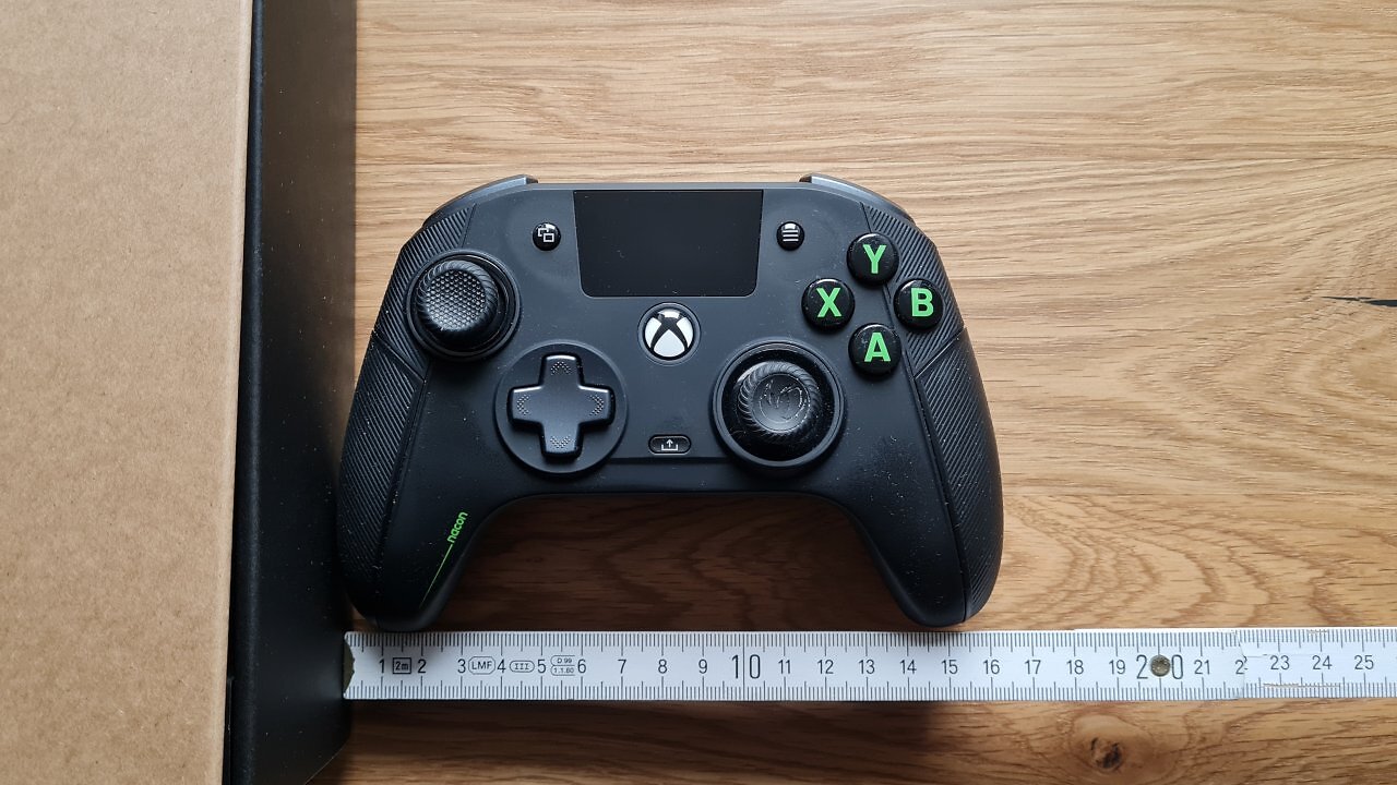 Ein schwarzer Controller auf einem Holztisch, darunter ein Meterstab, der die Breite zeigt.