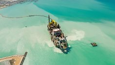 Luftaufnahme eines großen Baustellenschiffs auf türkisfarbenem Wasser in Küstennähe.