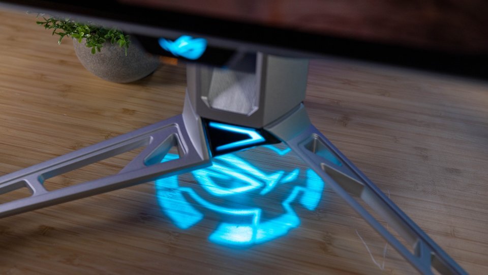 Der Standfuß des Asus ROG Swift OLED PG27AQWP-W ist nicht nur aus Leichtmetall gefertigt, sondern obendrein mit 'sportlich' wirkenden Aussparungen samt Verstrebungen versehen. Zum silbernen Farbton passt das nun in Türkis auf die Tischplatte projizierte Logo.